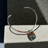 A silver-toned wire cuff bracelet with a small heart-shaped glass pendant featuring red, green, and white floral patterns, displayed on a light concrete block in direct sunlight.