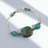 A silver-toned bangle bracelet featuring a large, organic-shaped green stone centerpiece and several smaller turquoise-blue beads, displayed on a light gray background with soft diagonal shadows.