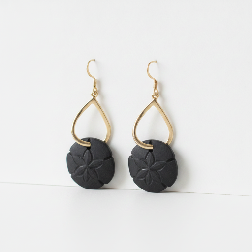 A pair of gold and black dangle earrings on a clean white background. Each earring features a gold teardrop-shaped wire frame suspending a round, textured black disc with a floral petal pattern.