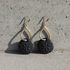 A pair of black circular drop earrings with an embossed floral pattern, suspended from gold teardrop-shaped hooks with fishhook wires, resting on a textured gray concrete surface.