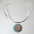 A silver-toned open-cuff necklace featuring a circular turquoise pendant with a red and white spiral design, shown against a plain white background.