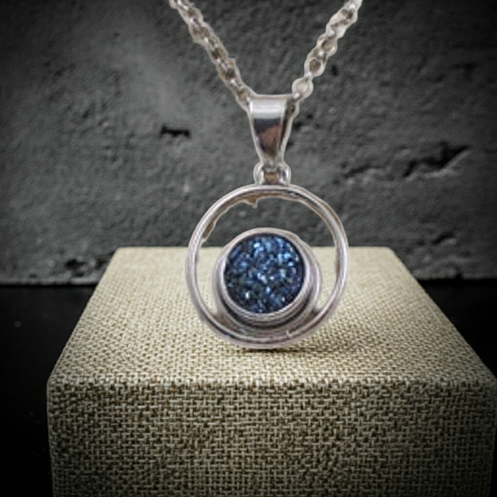 A silver pendant necklace with a circular frame and a blue glittery stone in the center, displayed on a textured beige block against a dark gray background.