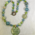 A handmade beaded necklace with a large green lotus pendant, featuring light green, teal blue, and beige beads, with a small gold-toned snake charm and textured round beads.