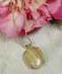 A silver-toned necklace with an oval pendant made from a polished, striped natural shell, resting on a textured white fabric next to pink flowers.