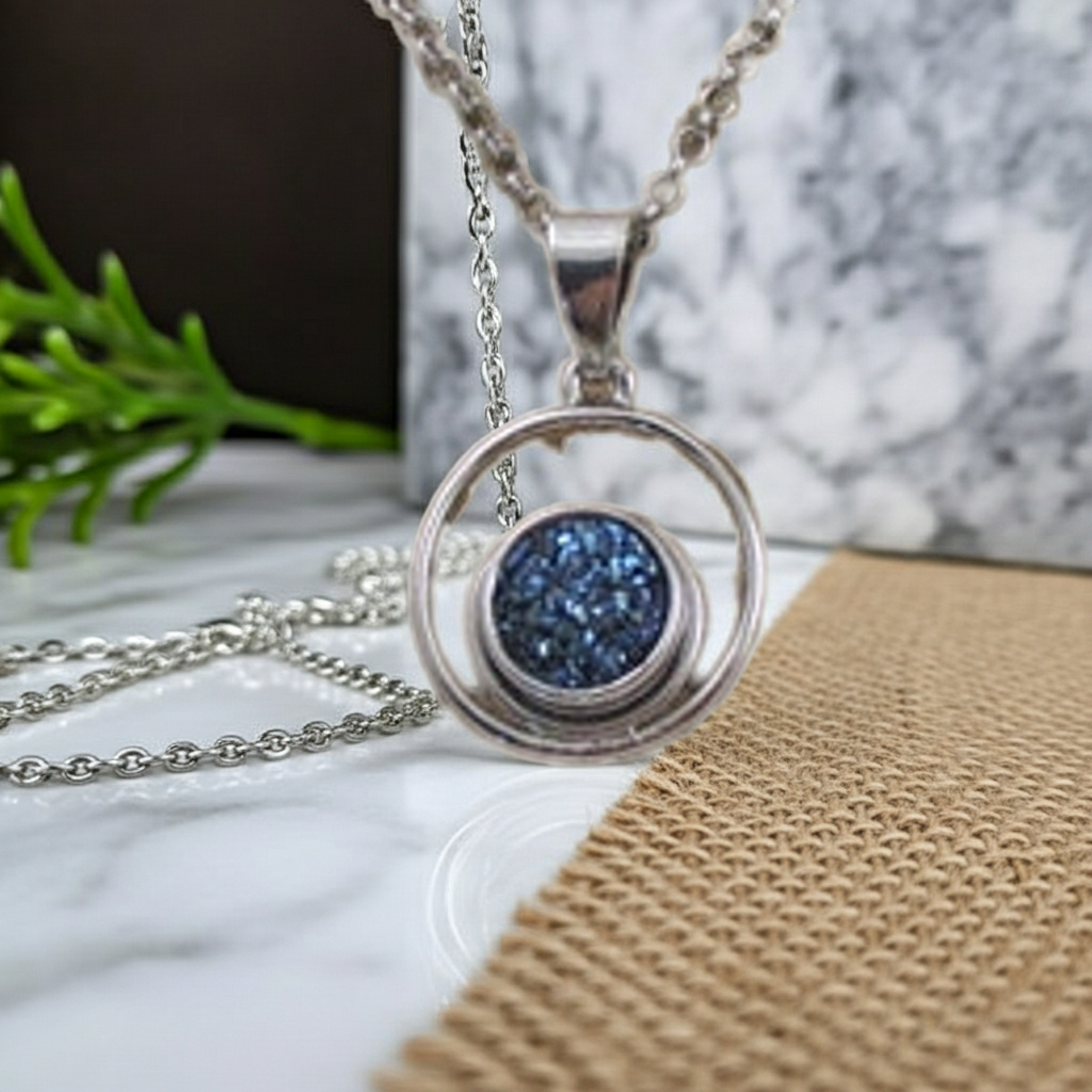 A silver-toned necklace with a circular pendant featuring a textured blue druzy stone, displayed on a marble surface with a burlap cloth and green sprig in the background.