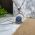 A silver-toned necklace with a circular pendant featuring a textured blue druzy stone, displayed on a marble surface with a burlap cloth and green sprig in the background.