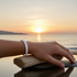 A white braided bracelet worn on a person's wrist, resting on driftwood at a beach during a golden sunset over the water.