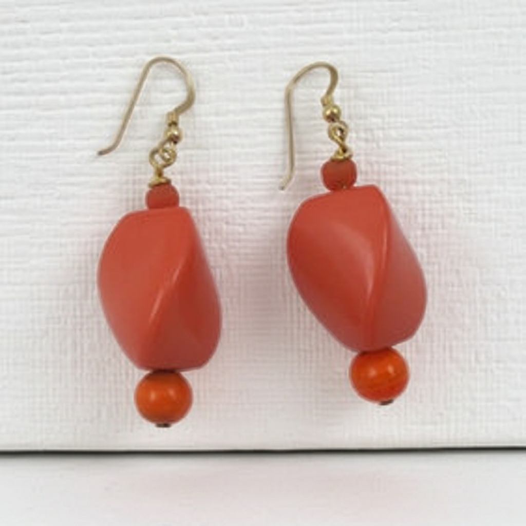A pair of coral drop earrings with gold-tone fishhook wires, featuring a large faceted coral bead, a small coral spacer bead, and a smaller round orange bead at the bottom, displayed on a textured white background.