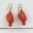 A pair of coral drop earrings with gold-tone fishhook wires, featuring a large faceted coral bead, a small coral spacer bead, and a smaller round orange bead at the bottom, displayed on a textured white background.
