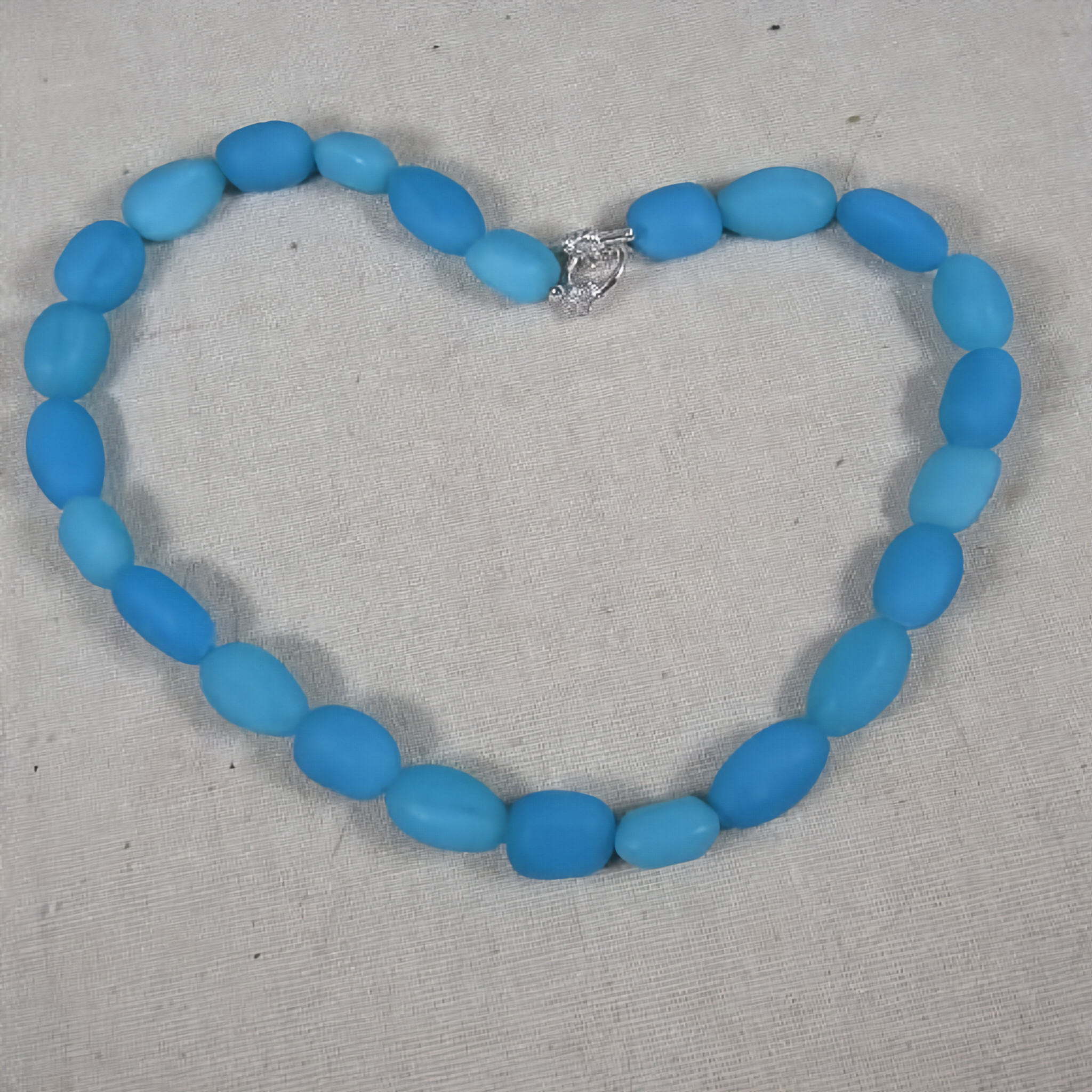 A bold blue opal beaded necklace made of smooth, rounded blue beads with a small silver clasp, displayed on a textured off-white fabric background.