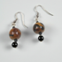 A pair of dangle earrings featuring polished brown and black tiger's eye beads, a small black spacer bead, and silver fishhook wires, shown against a light gray background.