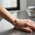 A close-up of a person's hand resting on a reflective gray desk, wearing a modern wrap bracelet with a bright orange leather strap, a silver-toned circular ring, and a small silver key charm.