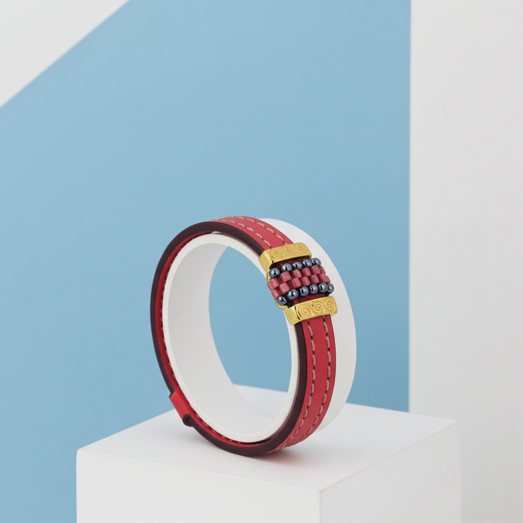 A red leather bracelet with a gold-tone buckle and a decorative center clasp featuring red, blue, and gold beads, displayed on a white geometric pedestal against a blue and white background.