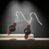 A pair of dangling earrings with clear fishhook wires, a small red bead, a larger dark brown bead, and a smaller red bead, displayed on a light surface against a dark textured background.