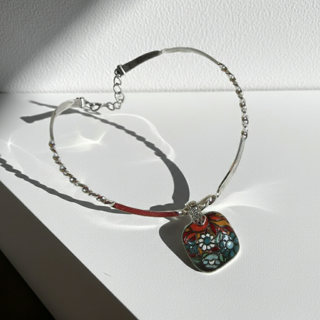 A silver-toned beaded chain bracelet with a decorative pendant, displayed on a white surface in direct sunlight. The pendant features a colorful floral pattern with red, blue, green, and white details.