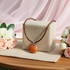 A round carved orange pendant necklace with a braided brown cord, displayed on a beige textured jewelry box. The box is styled on a wooden surface with pink and white roses against a soft pink fabric backdrop.